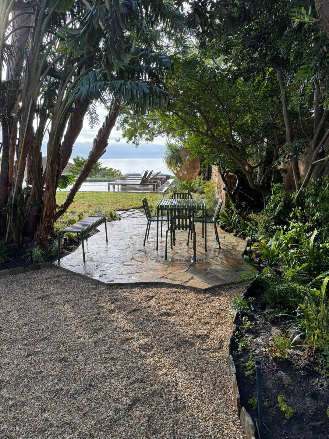 Raked gravel path under a canopy of mature trees leading to an outdoor dining table with a sea view