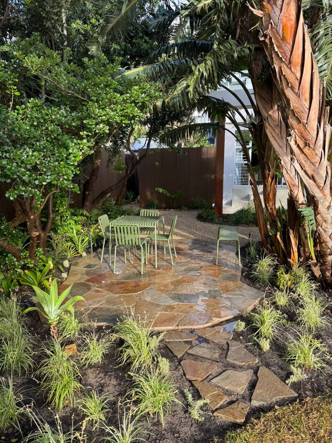 Natural flagstone path winding between fresh planting, green garden chairs and a corten steel fence
