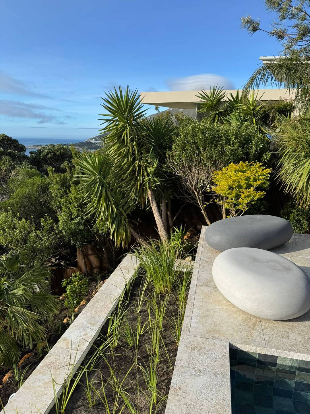 Raised travertine pool terrace with smooth sculptural stone seats, layered indigenous planting and a sea view