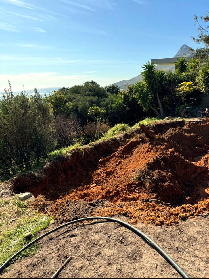 Steep garden slope stripped back to bare red earth during excavation, with the sea on the horizon
